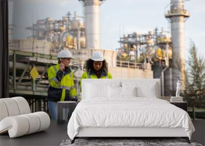 Two female engineers in high-visibility jackets and hard hats conducting a field inspection at an industrial plant with refinery structures in the background, ensuring workplace safety and efficiency Wall mural