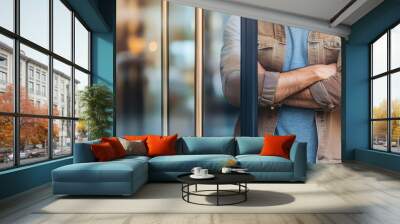 A stylish man with arms crossed stands confidently by a glass entrance, embodying modern elegance and urban lifestyle. Wall mural