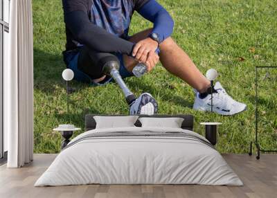 Close-up of Caucasian man with mechanical leg sitting on grass. Sportsman in blue shorts and white sneakers photographed in park. Sport, disability concept Wall mural