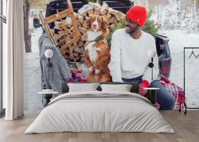 African American man sits with a dog in the trunk of a car in the winter forest. A man in a white sweatshirt. Wall mural