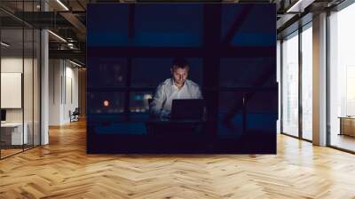 Businessman working on laptop in night office. Wall mural