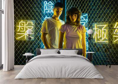 Young couple in white t-shirts stand against the background of a metal grid with neon symbols. Wall mural