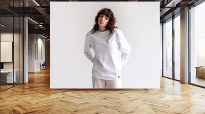 Young woman with curly hair in a white long sleeve t-shirt stands on a white background. Mock-up. Wall mural