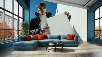 Man in Bold Winter Fashion with Black Outerwear Over White Sweater and Sweatpants Holding Large Textured White Magazine, Set Against a Sunlit Background with Vibrant White Wall and Blue Sky Wall mural