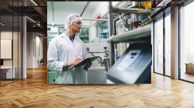 A man in a white lab coat is writing on a clipboard in front of a machine. Concept of focus and concentration as the man takes notes while working in a laboratory setting Wall mural