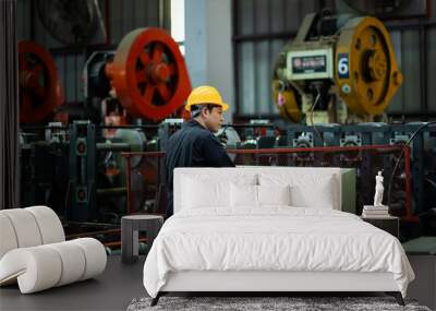 A man in a yellow helmet is working on a machine in a factory. The machine is large and red, and the man is focused on it. The atmosphere of the image is industrial and serious Wall mural