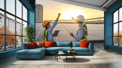 Two construction workers are standing on a roof, one of them is holding a tablet. They are both wearing hard hats and safety vests. Electrical engineer installs solar panels in power station Wall mural