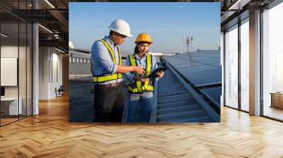 Two people are standing on a roof, one of them is pointing at a tablet. They are wearing safety vests and hard hats. Electrical engineer installs solar panels in power station Wall mural