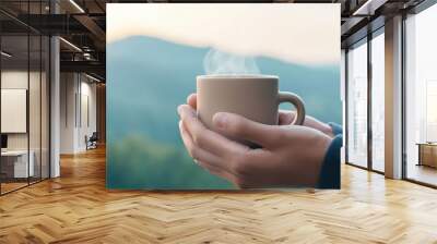 Person holding a steaming cup of coffee against a mountain backdrop. Wall mural