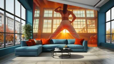 A woman exercises in a sunlit gym, flexing her muscles and embracing the morning light through large windows for a powerful workout session. Wall mural