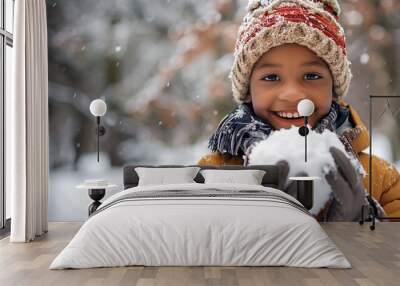 A cute and happy African American black boy dressing in brown winter clothes, holding a snowball on a snowy background with copy space for text. Wall mural