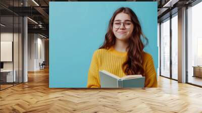 An imaginative white teenage girl wearing glasses is holding a green book and looking up on a plain blue background with copy-space for text. Wall mural