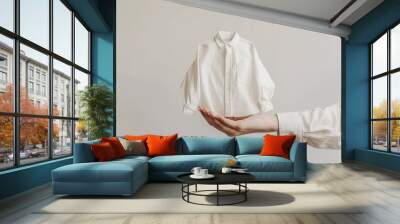 Mockup of tiny clothes. A cute white mini long sleeve shirt is held by a woman hands on a plain white background with copy-space for text suit for a fashion advertisement. Wall mural