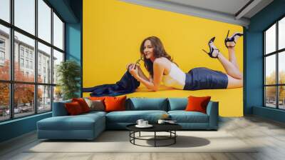 a young lady, brunette, dressed elegantly and stretched on the belly on a yellow background, with glasses in hand Wall mural