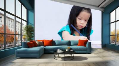 Asian adorable girl is doing homework. Children paint with wooden colors on a coloring book. White paper. Child learn art. On a white wall background. Kid is 4 years old. Home school. Stay at home. Wall mural