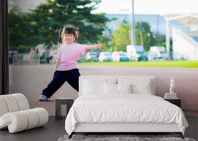 Asian girl is standing in stadium for exercise. Child stand with their legs and arms extended. Happy kid is 4 year old. Lady wearing pink shirt, smiling sweet. Concept of children playing light sport. Wall mural