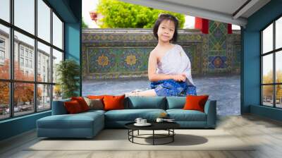 Cute girl wears Thai dress with silver sash and blue loincloth trimmed with gold. Asian kid visits temple in Thailand to see art and culture with her family. Child squats sit on the floor and smiles. Wall mural