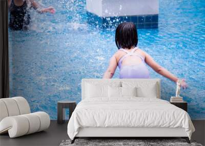 Rear view of Asian cute baby girl is splashing water from a pool on her mother. Children play in the water and have fun with the family. Child wear purple swimsuits. Kid aged 5-6 years old. In summer. Wall mural