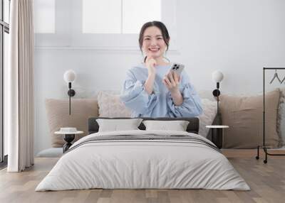 Young woman thinking with her phone in a beautiful living room, looking at the screen, smiling and finding something to do, front, looking at the camera. Wall mural