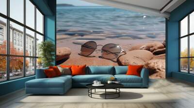 A pair of sunglasses is laying on the beach. The beach is rocky and the water is calm Wall mural