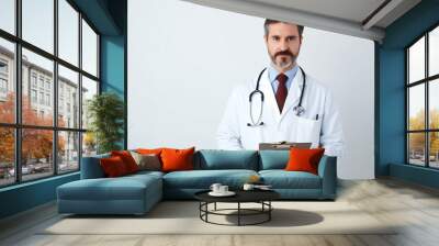 Professional doctor man in medical coat with clipboard in hands posing over light white wall background, middle aged physician with stethoscope looking at camera Wall mural