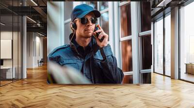 Security guard man in uniform cap, jacket and sunglasses is standing by window inside house and talking on portable wireless two way walkie talkie transceiver radio set device. Property safety concept Wall mural