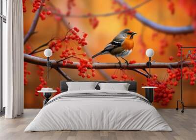 A small bird perches on a branch laden with red berries against a vibrant autumn backdrop Wall mural