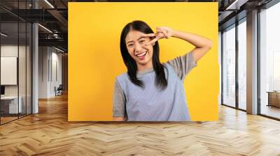 Young asian woman in casual clothes smiling and showing v sign gesture to covering eye with cheerful Wall mural