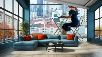 Business remotely. A girl works at her laptop on a balcony in the city center with a landscape view Wall mural