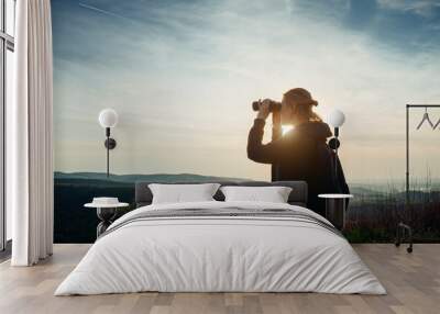 A young adult female hiker and traveler looks through binoculars in the mountains in the magical evening light of a sunset Wall mural