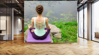 Thin brunette girl plays sports and performs beautiful and sophisticated yoga poses in a summer park. Green lush forest and the river on the background. Woman doing exercises on a yoga mat Wall mural