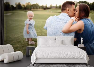 A couple hugging and kissing while sitting on the grass, and their beautiful daughter runs nearby Wall mural