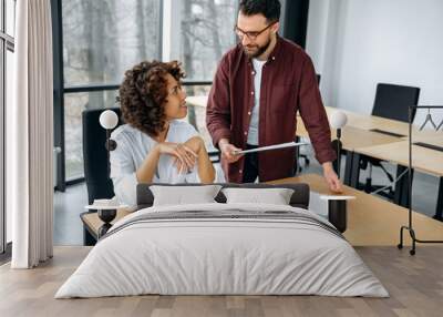 African american woman and caucasian man, company colleagues, work together on a project in a modern office, advise each other, share ideas and solutions, plan a product promotion strategy, smile Wall mural