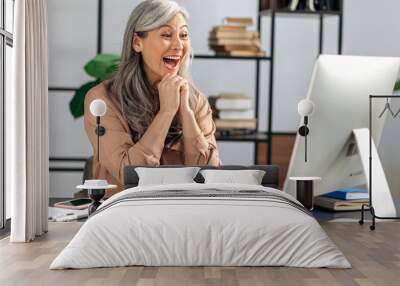 Amazed senior gray-haired happy Asian woman, freelancer, manager, looks at the computer screen, made good profit or news, made profitable deal, gestures with her hands, joyful expression on her face Wall mural