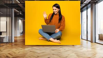 Attractive caucasian brunette girl stands on an isolated orange background holds a laptop looks directly into the camera with friendly smile, sitting on isolated orange background Wall mural