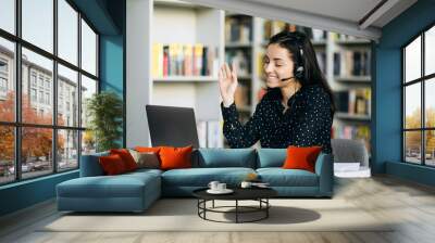 Attractive female employee sit at the work desk, having a video conference with colleagues or clients. Caucasian young woman wearing headset, works as call center operator, communication concept Wall mural