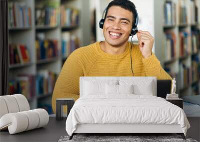 Attractive hispanic young man, call center worker, student, or successful business man, in a headset is using a laptop while working online sitting at his desk and looking at the camera with smile Wall mural