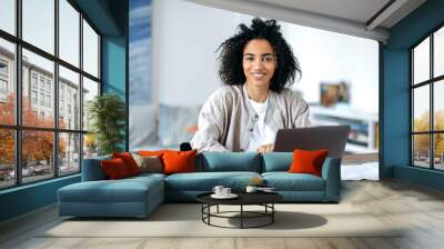 Beautiful friendly confident young african american woman in stylish casual clothes, freelancer, manager or student sitting at laptop, working or studying at home, looking at camera, smiling Wall mural