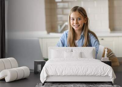 Caucasian pretty blonde schoolgirl is learning at home using laptop, sitting at the work desk, looks at the camera and smiling. Education during quarantine Wall mural