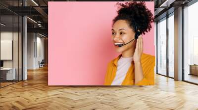 Close-up of positive african american curly haired young woman with headset, help desk worker, call center operator, consultant, talking with client, stand on isolated pink background, smiles friendly Wall mural