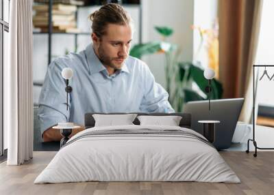 Concentrated caucasian guy freelancer or student uses laptop, distant learners or work. Focused attractive modern man in stylish casual wear sits at the desk in a creative or home office, browsing Wall mural