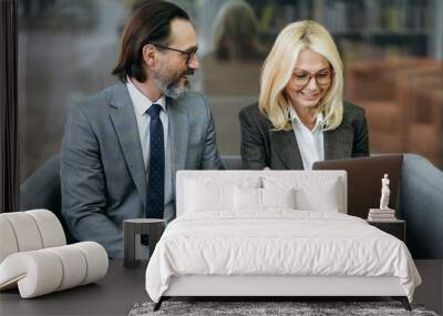 Confident middle aged business partners sitting on the sofa, discussing project, smiling. Male manager and elegant female employee using laptop, searching ideas, browsing internet, teamwork concept Wall mural