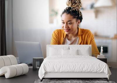 Focused cute stylish african american female student with afro dreadlocks, studying remotely from home, using a laptop, taking notes on notepad during online lesson, e-learning concept, smiling Wall mural