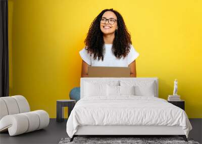 Full length photo of positive joyful creative young brazilian or hispanic woman with glasses, freelancer or designer, sit on isolated orange background with laptop, looks to the side, dreaming, smiles Wall mural