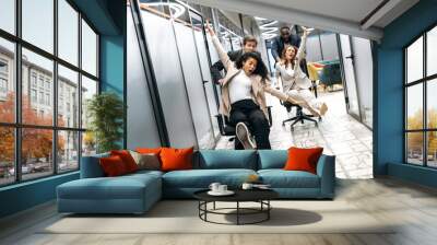 Funny young adult business people making a race on chairs in modern office. Multiracial friendly colleagues spending together their work break, resting from brainstorm Wall mural