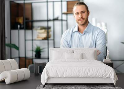 Handsome confident attractive caucasian stylishly dressed guy, designer, freelancer or employee, standing near his desk, with arms crossed, looking at the camera and smiling friendly Wall mural