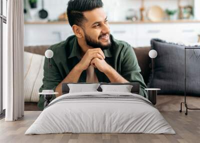 Handsome positive confident attractive indian or arabian man, in casual stylish clothes, sitting at home in cozy living room on the background of the kitchen, looks to the side, smiling Wall mural