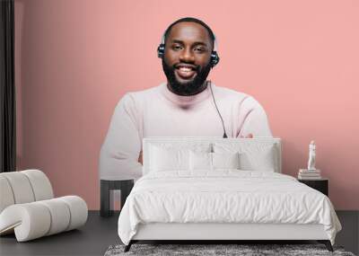 Handsome young african american man worker of a call-centre with headphones is standing on a pink isolated background looks at the camera and smiling crossed arms in front of him Wall mural