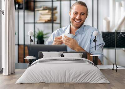 Happy friendly confident caucasian guy, employee or freelancer, holding a cup of coffee while sitting at his desk at home or office, looking at the camera with a friendly smile Wall mural