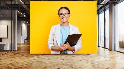 Happy positive lovely mixed race female employee, real estate agent, recruitment specialist, hr manager, holding documents, stands on isolated orange background, looking at camera, smiling friendly Wall mural